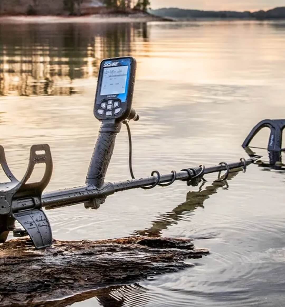 Detector de metales Nokta Double Score mojado y apoyado en un tronco flotante dentro de un lago.