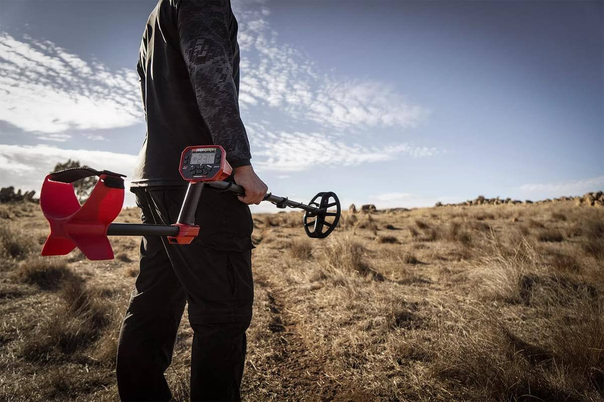 Vista trasera de un hombre sosteniendo el Detector de metales Minelab Vanquish 540 en un campo seco bajo el sol.