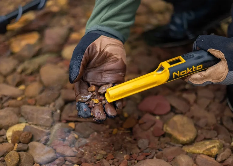 Operador con guantes de cuero utilizando el localizador Nokta AccuPoint en un lecho de río pedregoso.