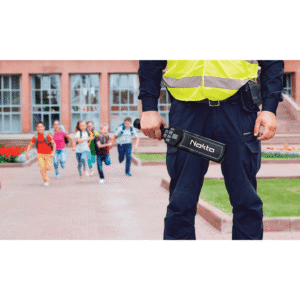 Vigilante con chaleco reflectivo sosteniendo el detector de metales manual Nokta NMS20 en la entrada de un colegio con niños corriendo de fondo.