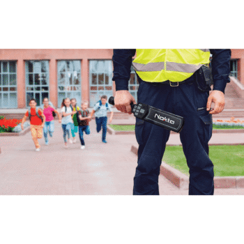 Vigilante con chaleco reflectivo sosteniendo el detector de metales manual Nokta NMS20 en la entrada de un colegio con niños corriendo de fondo.
