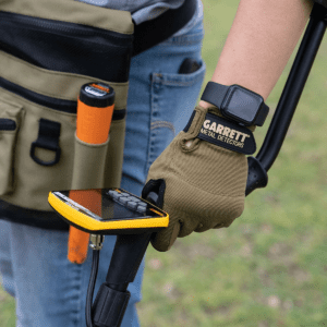 Operador usando un guante táctico verde oliva Garrett para sujetar la empuñadura de un detector de metales con pantalla amarilla en un campo abierto.