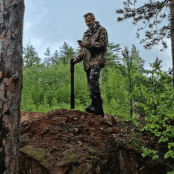 Usuario profesional de camuflaje utilizando el escáner de suelo OKM Fusion Ultra Light y un smartphone en un entorno forestal.